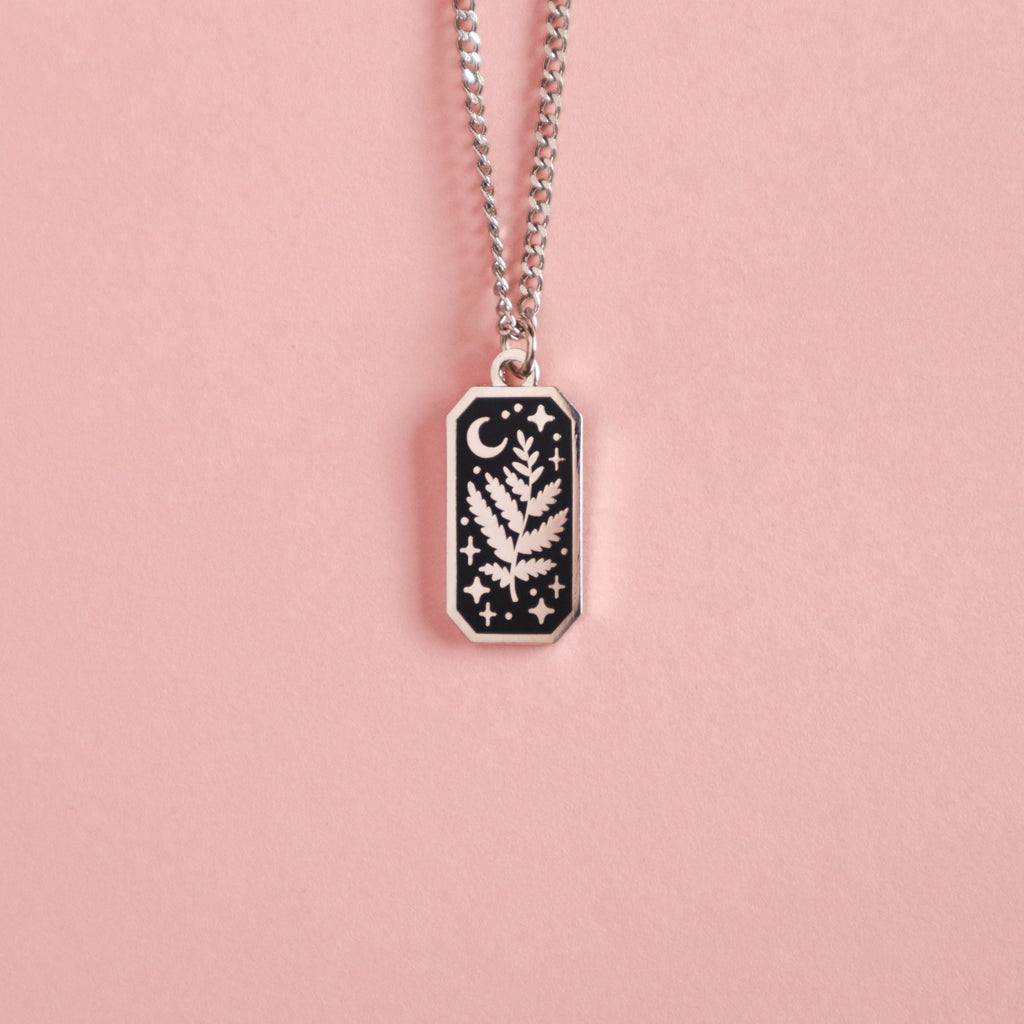 A photograph of a rhodium plated necklace. The necklaces are rectangular and feature a leaf, moon and stars and is taken on a pink background.
