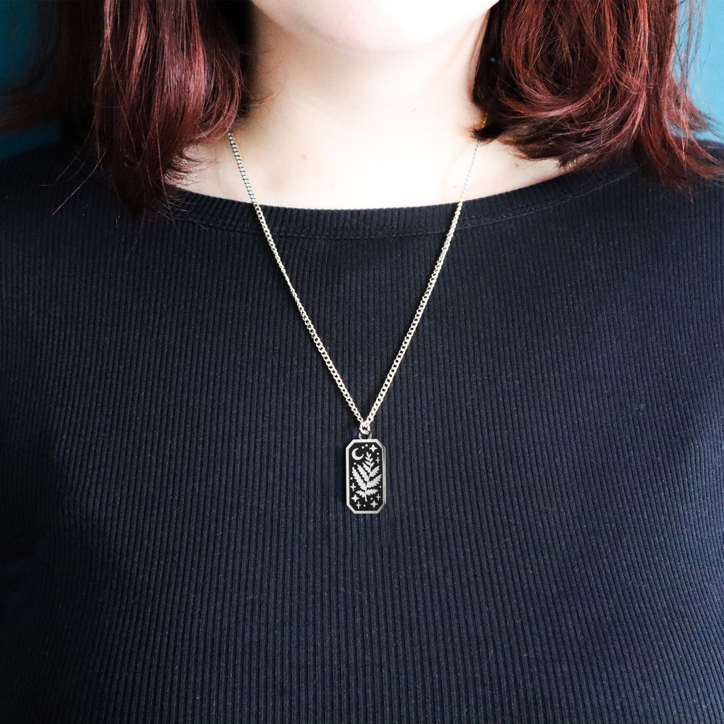 A photograph of a rhodium plated necklace on a model with red hair and a black sweatshirt. The necklace is rectangular and features a leaf, moon and stars. 