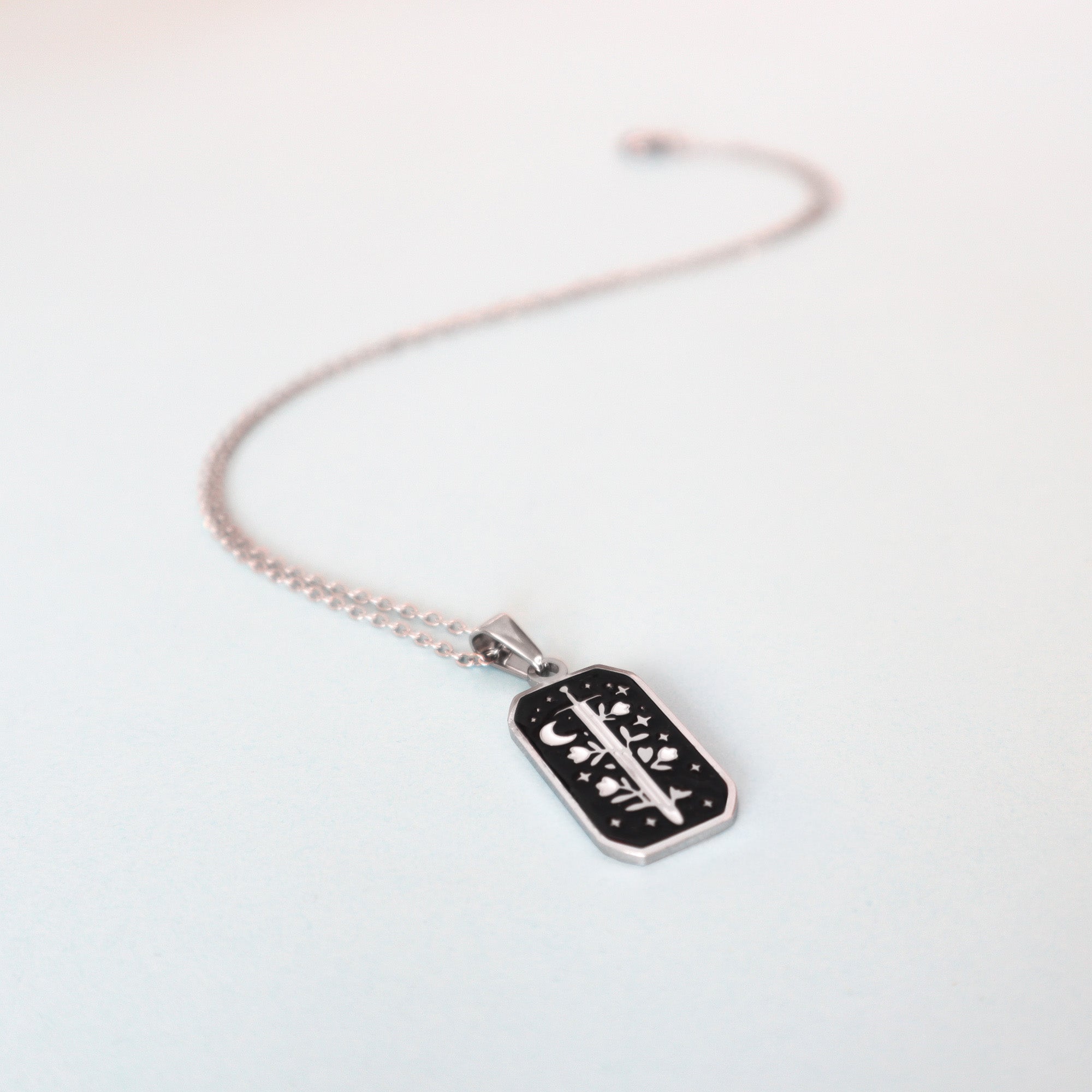 The Warrior Enamel Necklace by Papio Press features a silver chain and a rectangular black enamel pendant with a sword, crescent moon, and stars—an ideal piece of fantasy jewelry shown on a light surface.