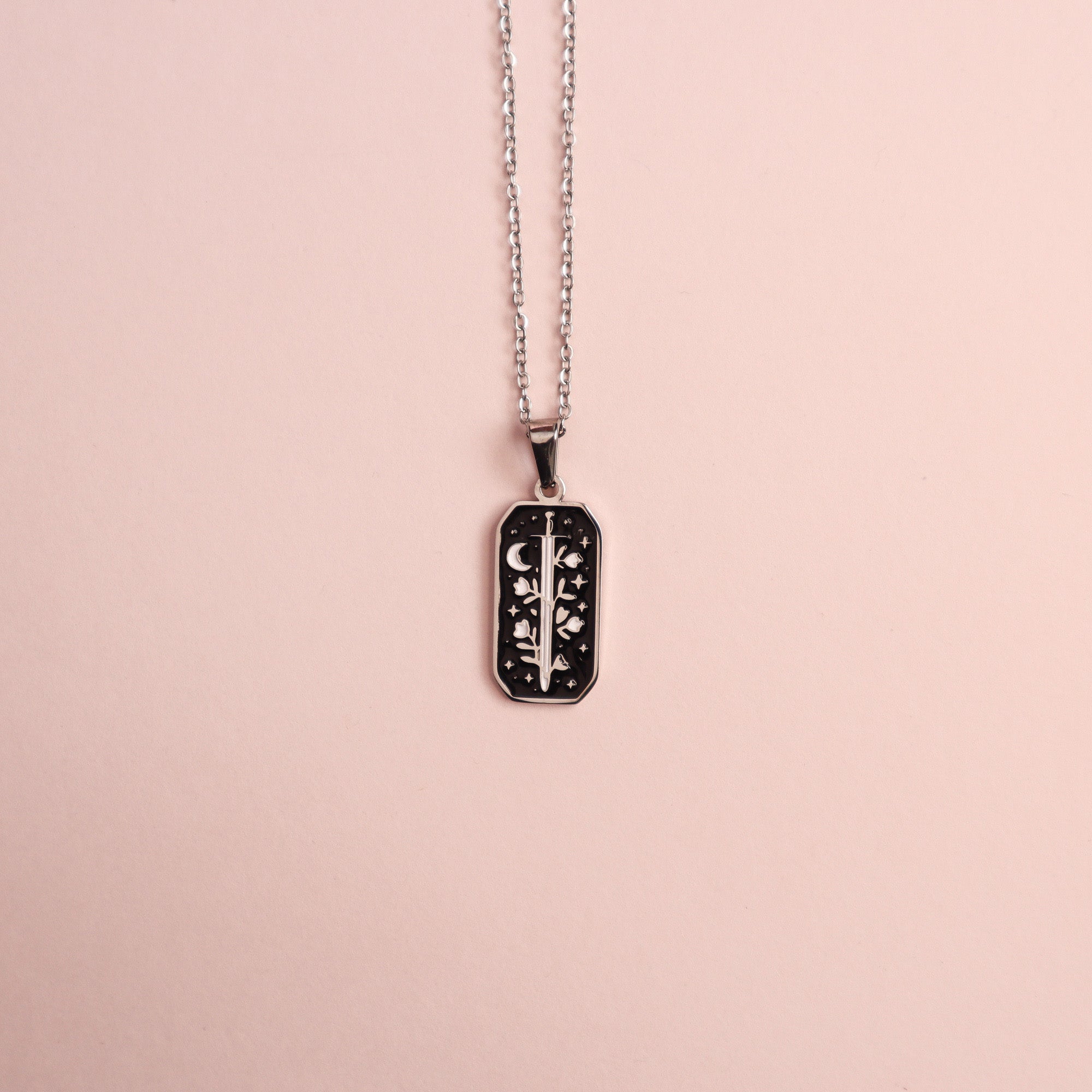 The Warrior Enamel Necklace by Papio Press, featuring a rectangular pendant with a detailed floral design, is showcased on a pale pink background.