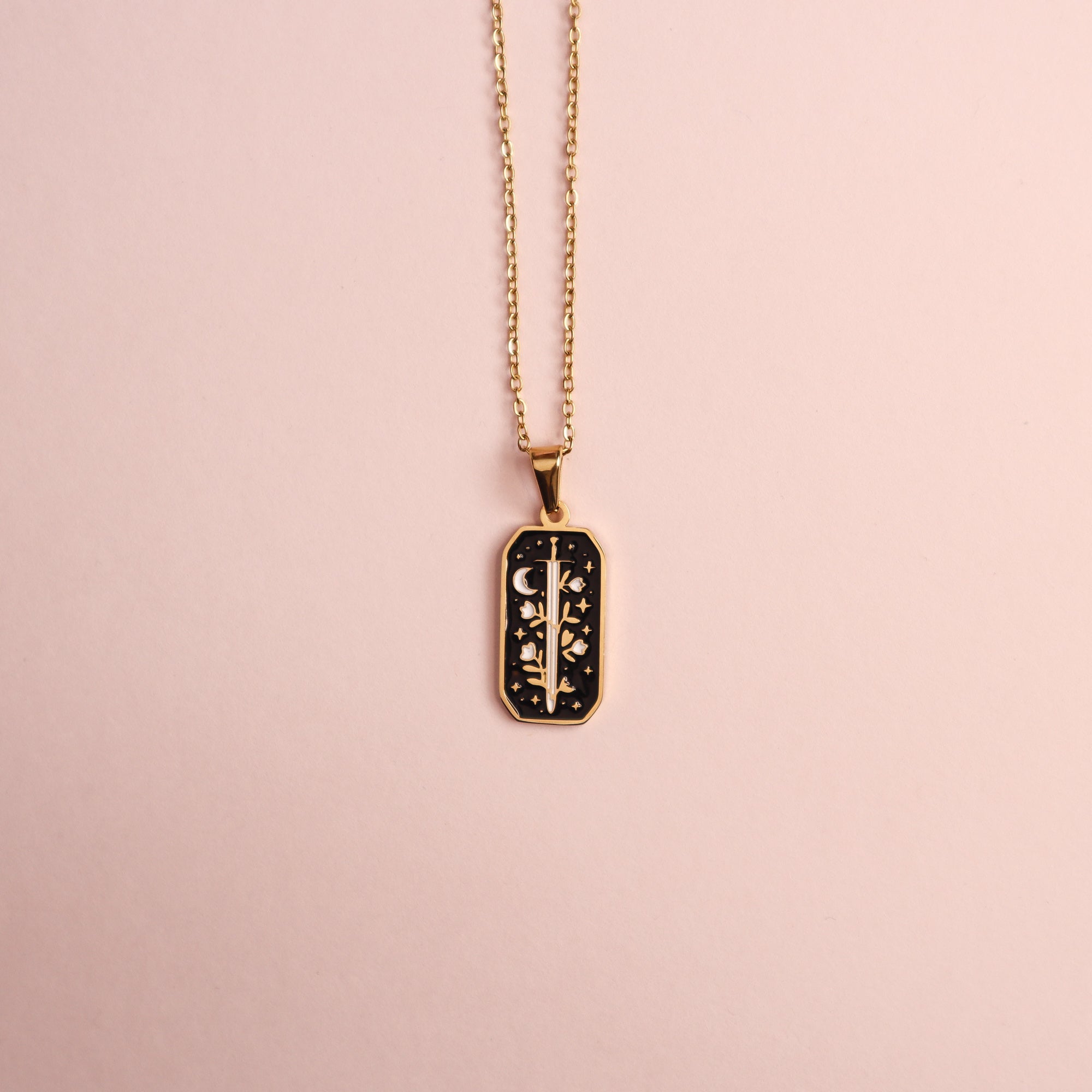 The Warrior Enamel Necklace by Papio Press features a gold chain with a rectangular black pendant, showcasing a symmetrical floral and crescent moon design on a light pink background.