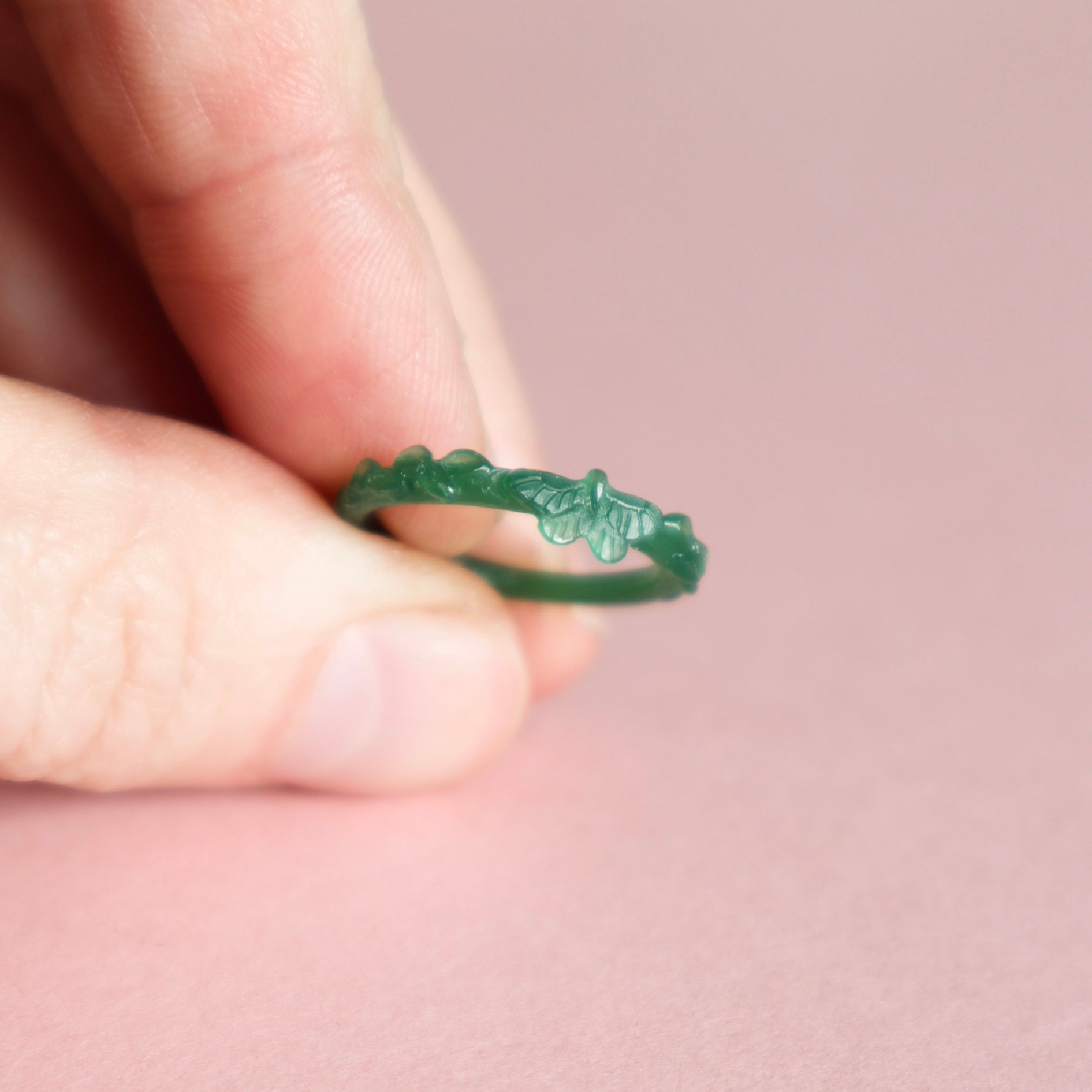 Green wax butterfly ring held between fingers against a pink background.