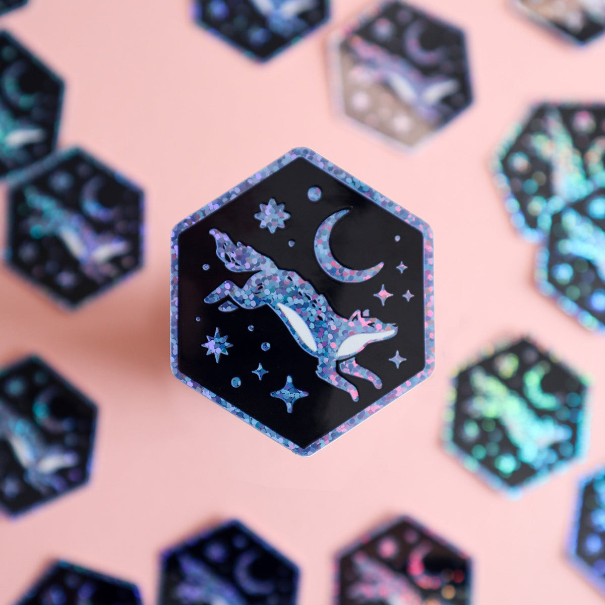 A hexagonal sticker featuring a glittery blue and purple leaping wolf under a crescent moon and stars on a black background, displayed on a soft pink surface with other similar stickers blurred in the background.