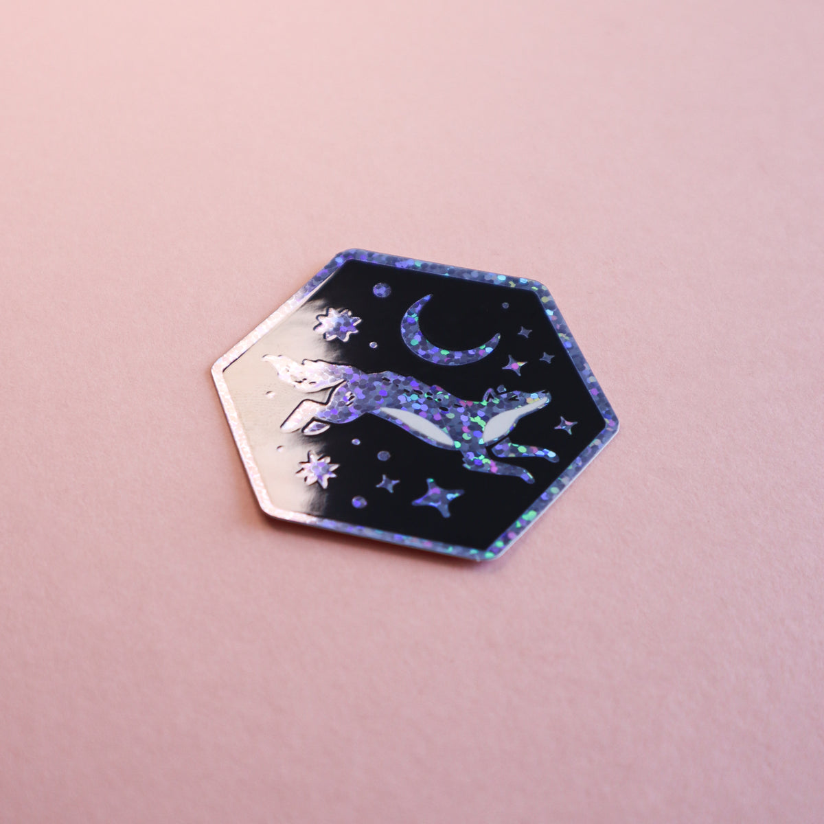 A hexagonal sticker featuring a glittery purple and blue wolf jumping over a crescent moon and stars on a black background, displayed on a soft pink surface.
