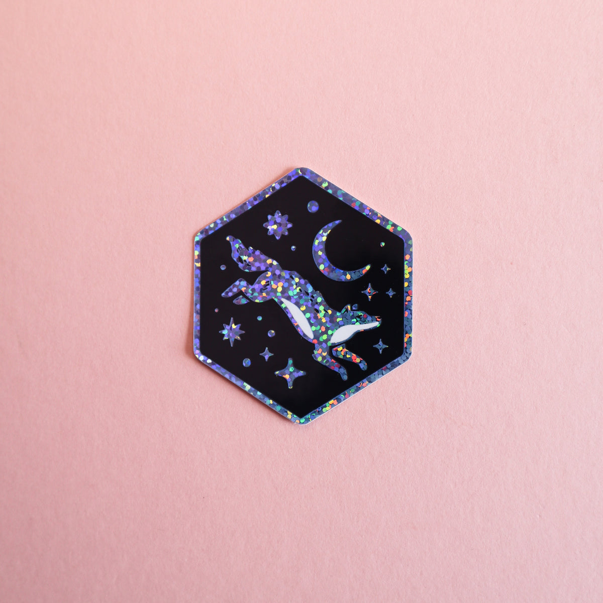 A hexagonal sticker featuring a black background with a colourful holographic glittery wolf jumping towards a crescent moon and stars, set against a solid pink background.