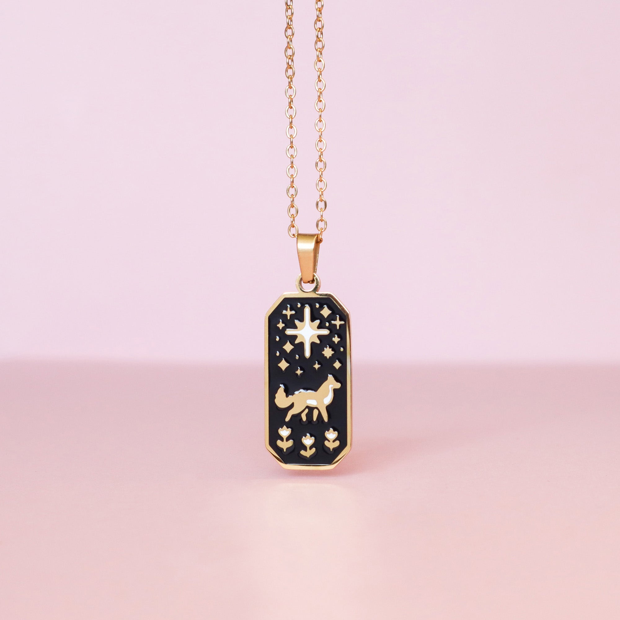 Papio Press Wolf & Star Enamel Necklace with a rectangular pendant features a wolf, stars, and floral designs on black enamel. Displayed against a soft pink background.