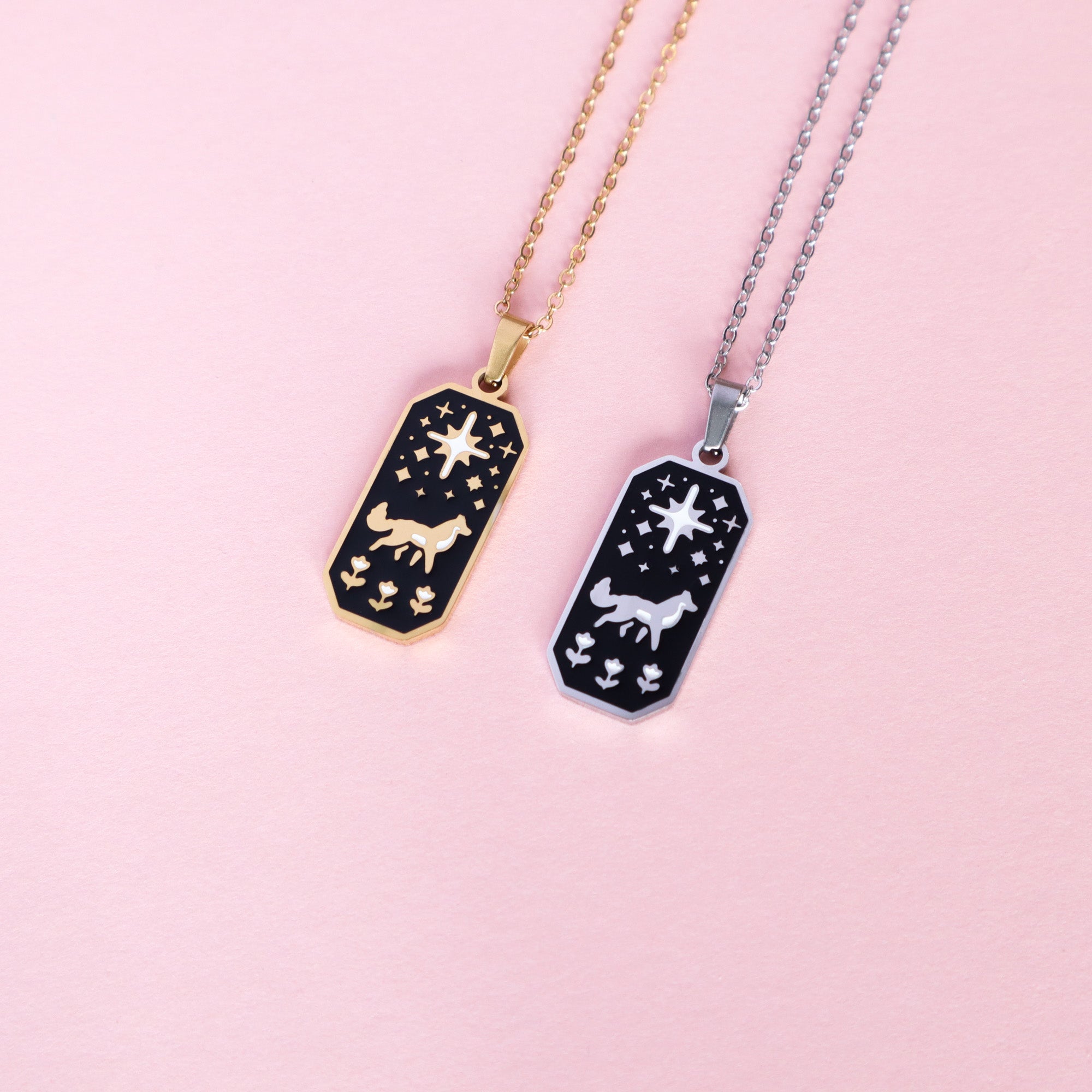 The Wolf & Star Enamel Necklaces by Papio Press feature rectangular gold and silver pendants with a wolf, stars, and plants on black enamel. These stainless steel pieces are shown on a light pink surface.