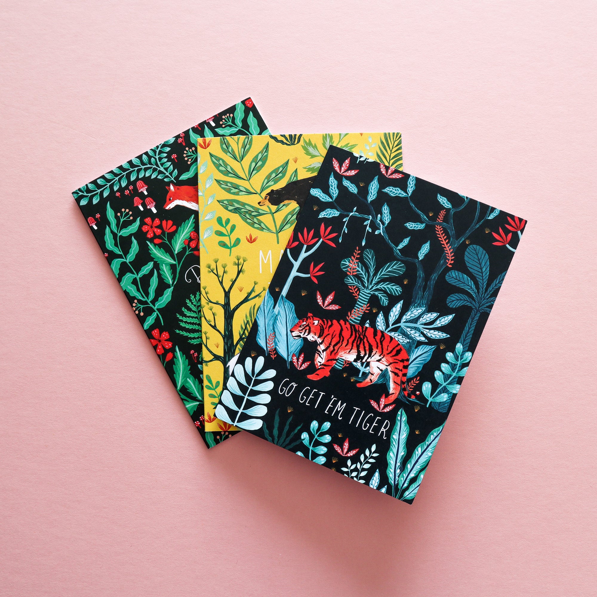 A pack of 12 Animal Greetings Cards by Papio Press, featuring vibrant animal and botanical illustrations—including a tiger—is displayed on a pink surface. The cards are made from recycled card and come with bold, eye-catching designs.