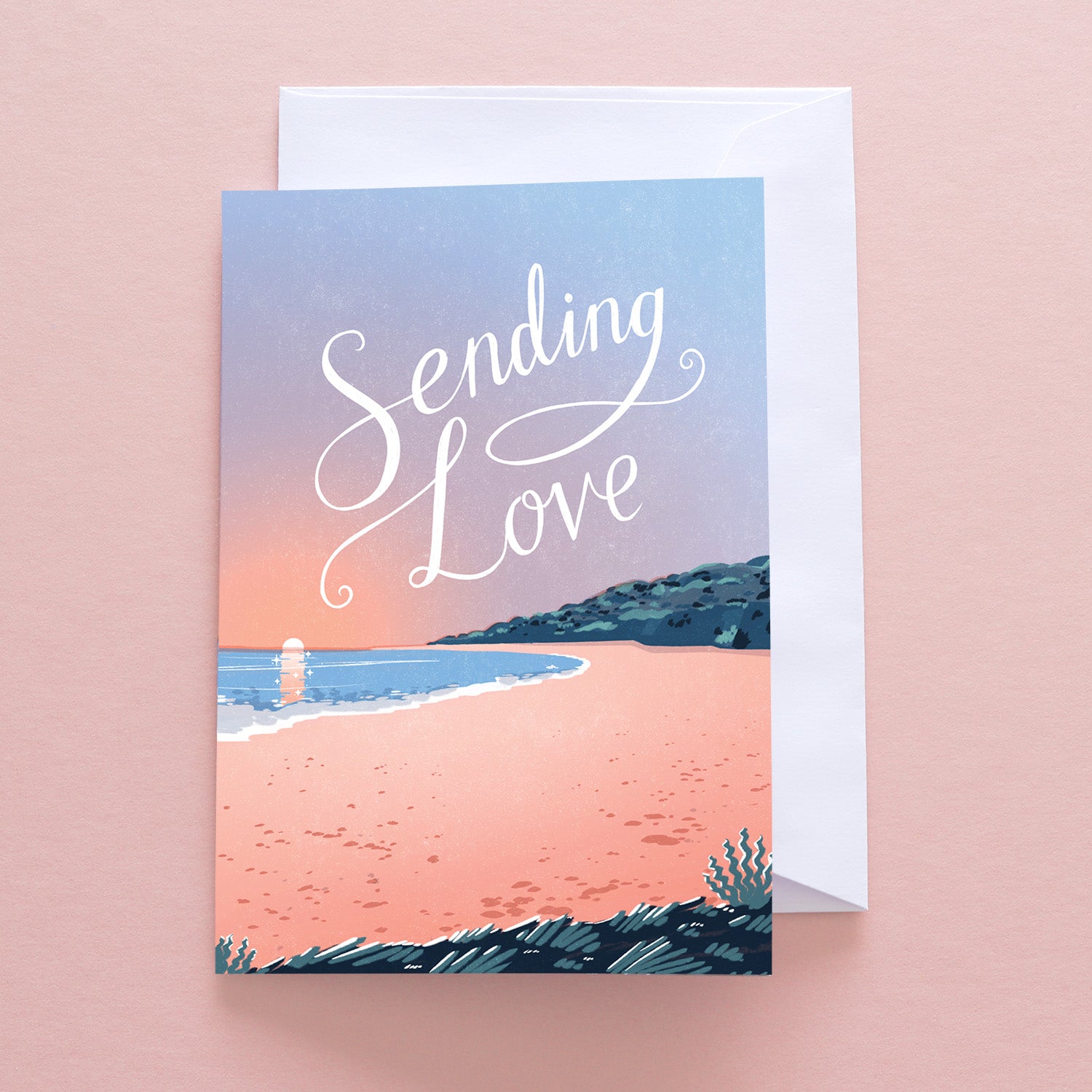 The Papio Press Sending Love Beach Greetings Card sits on a pink surface, displaying a peaceful sunset and “Sending Love” in white script. A recycled envelope is partially visible behind the card.