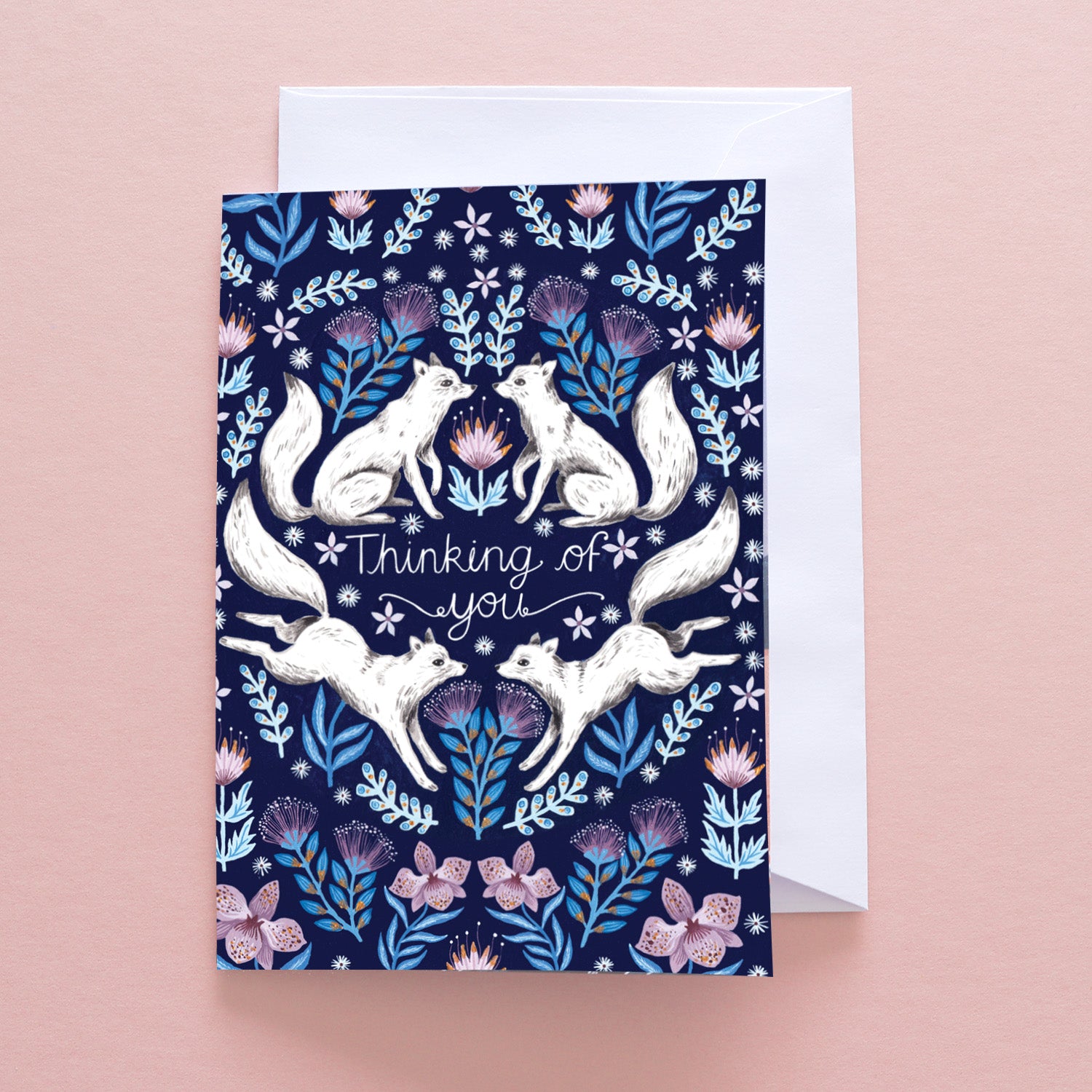 The Papio Press Thinking Of You Arctic Foxes Greetings Card features white foxes with pink and blue flowers on navy recycled stock. Displayed with a white envelope, the front reads “Thinking of you.”.