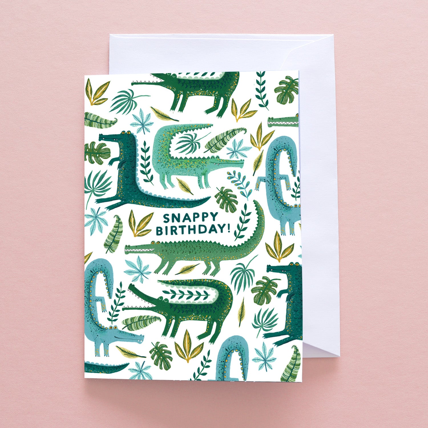 The Have A Snappy Birthday Greetings Card by Papio Press features green and blue cartoon alligators, leaves, and plants with SNAPPY BIRTHDAY! on a pale pink background. Comes with a white recycled stock envelope.