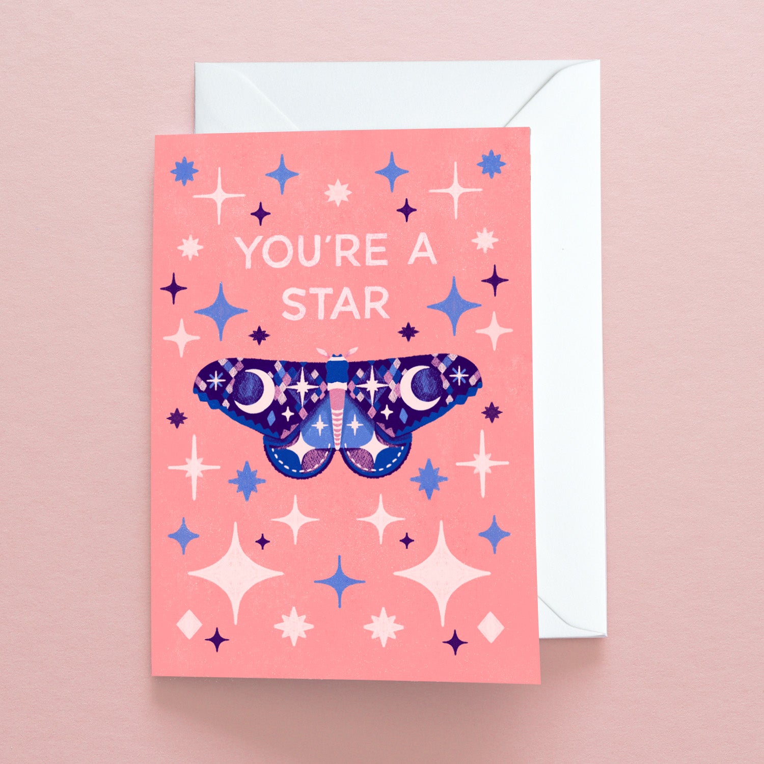 The Youre a Star Moth Greetings Card by Papio Press features YOURE A STAR in white, a blue-purple moth with stars and sparkles, is printed on recycled stock, includes an envelope, and comes on a pink background.