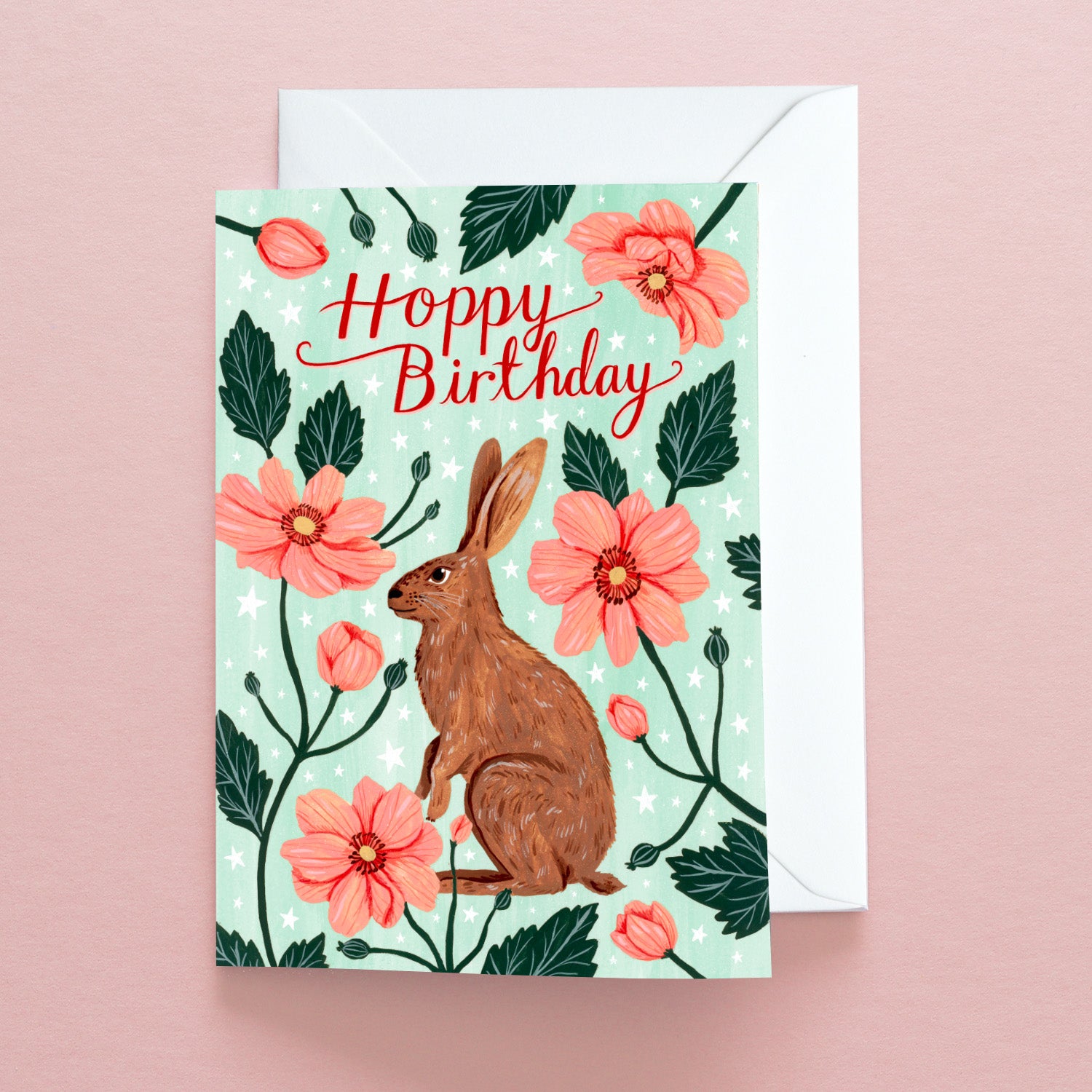 The Hoppy Birthday Greetings Card by Papio Press features a brown rabbit and pink flowers on light green recycled stock, with Hoppy Birthday in red script. It comes with a white envelope and is displayed on a pale pink surface.