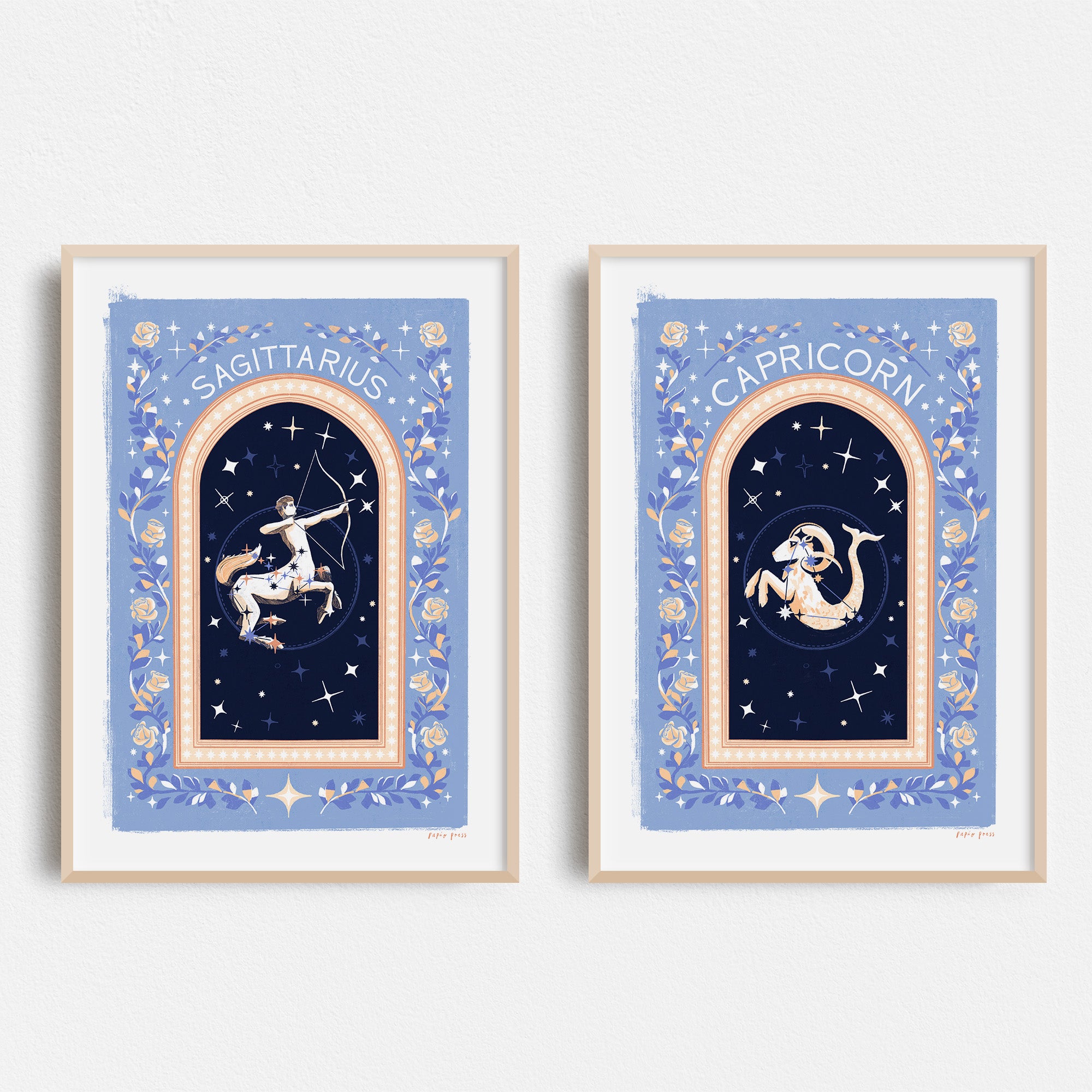 Two Choose your own Constellation Print // Star Signs art prints by Papio Press hang on a white wall, unframed, each with a blue starry background and floral borders, depicting Sagittarius and Capricorn.