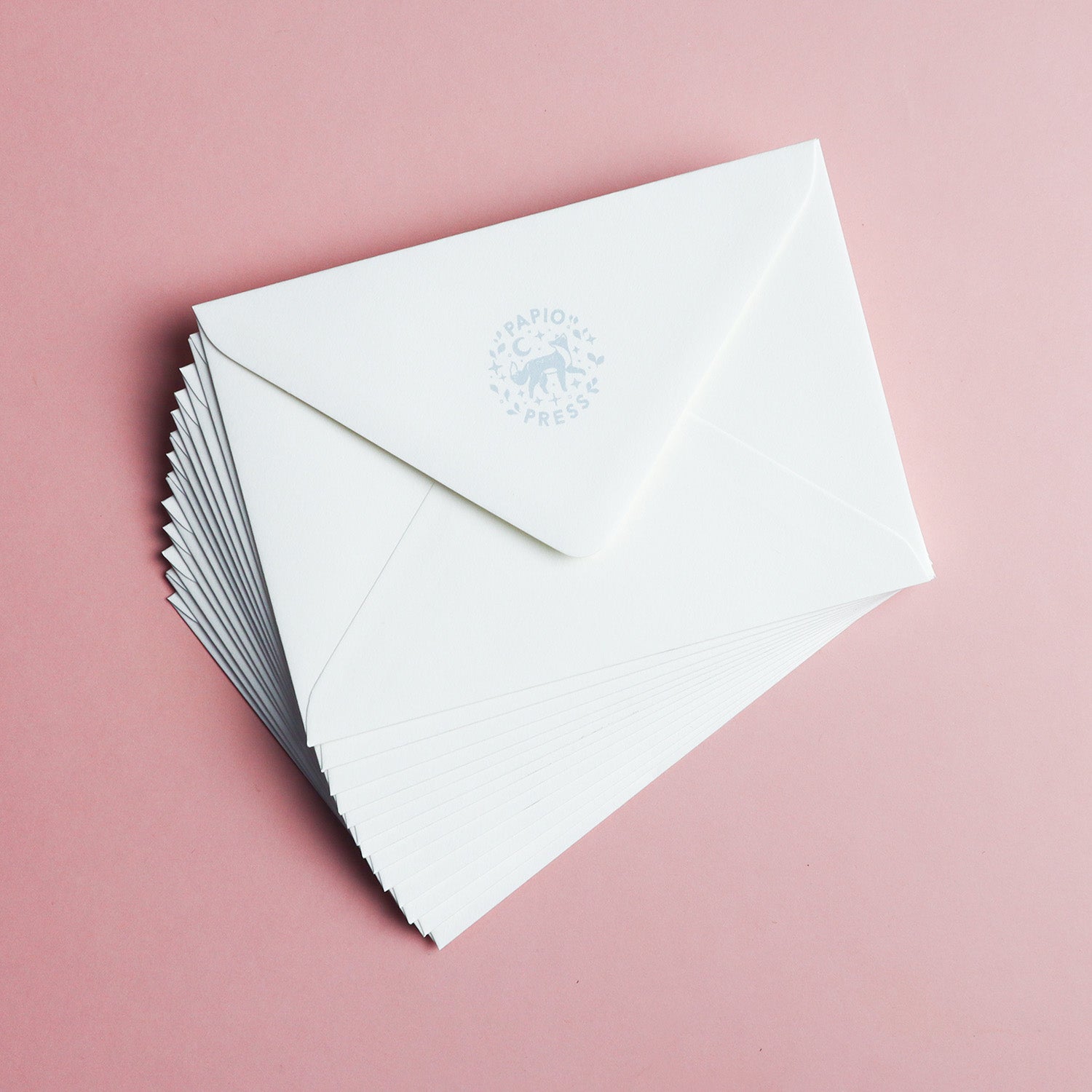 A stack of white envelopes made from recycled stock is fanned out on a pink background. The top envelope shows the Papio Press logo, and it pairs with the Have a Magical Birthday Jackalope Greetings Card by Papio Press.