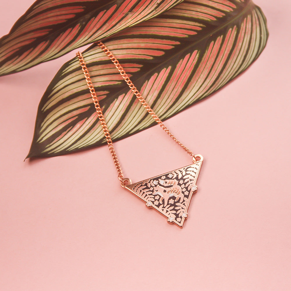 A triangular rose gold enamel necklace featuring a fox surrounded by florals, resting on a leaf on a pink background.