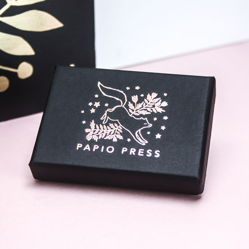Papio Press necklace packaging. A black paper box, featuring rose gold foiling of a leaping fox surrounded by florals. 
