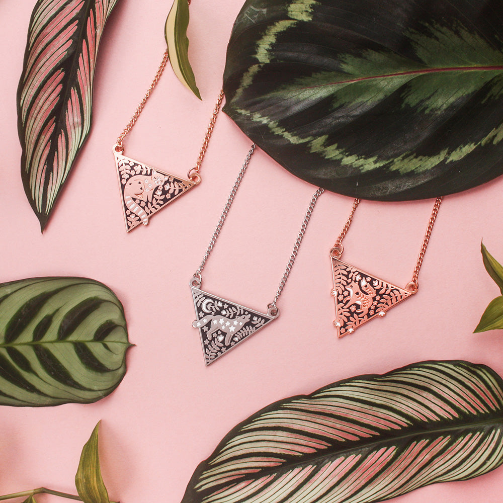 A collection of triangular necklaces surrounded by leaves, resting on a pink background. 