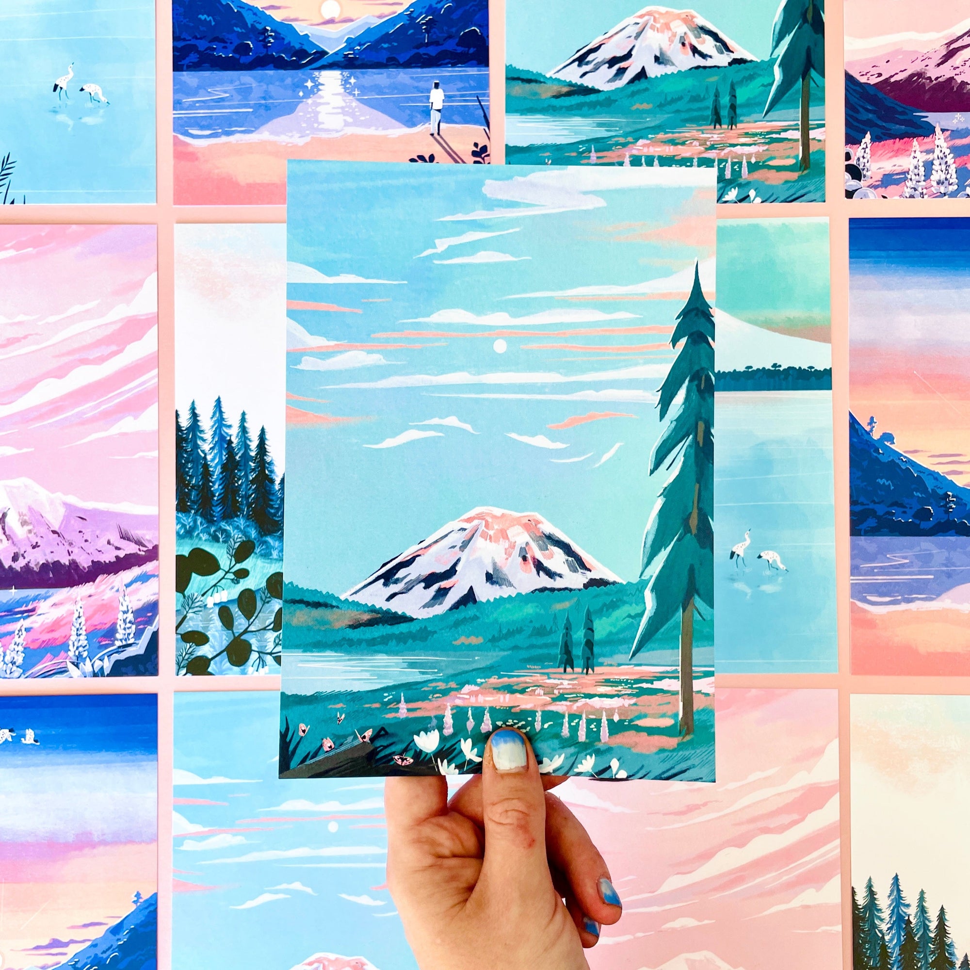 A hand with pastel blue nails holds a Papio Press A5 postcard from the “5 Large Papio Press A5 Postcards with Mountain Illustrations” set, featuring snowy peaks and pines, aligned with other vibrant recycled card artworks in the background.