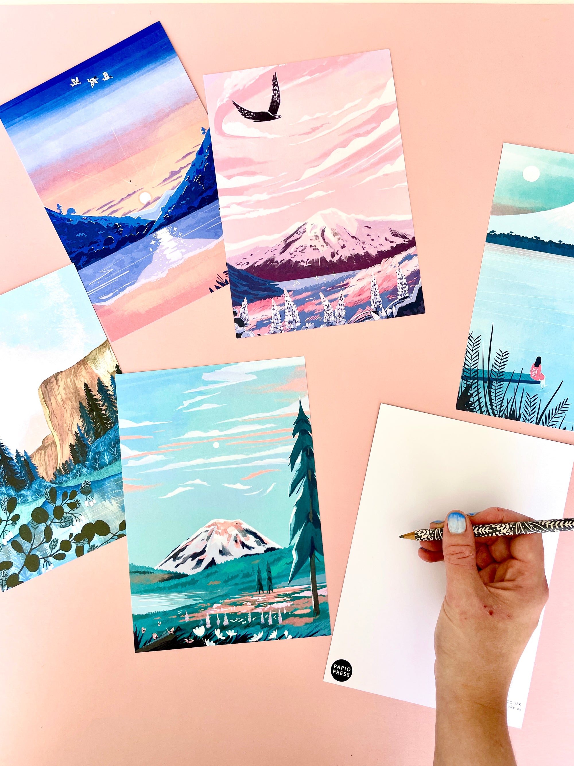 Five large Papio Press A5 postcards with mountain illustrations are shown on a peach surface. A hand with blue nails writes on the blank back. Each eco-friendly postcard is printed on recycled card by Papio Press.